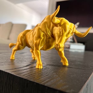 May include: A gold-colored bull figurine, standing on a black wooden surface. The bull is facing the right side of the image, with its head slightly tilted down. The figurine is detailed with a textured surface, giving it a realistic appearance.