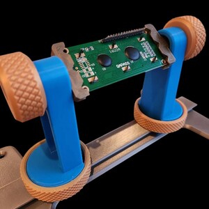 May include: A blue and tan circuit board holder with a green circuit board. The holder has a silver base and tan knobs. The circuit board has two black buttons and the text "QADASS".