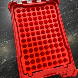 May include: A red, open storage case with a clear lid. The interior features a grid of circular holes. The text "12 Gauge" is visible on the case. The case is designed to hold small, cylindrical objects.