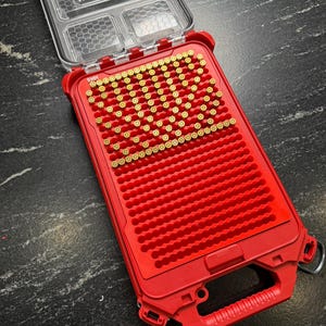 May include: A red plastic storage case with a clear lid, open to reveal brass bullet casings. The case has a handle and is on a dark surface. The lid has several small compartments. The case is designed for ammunition.