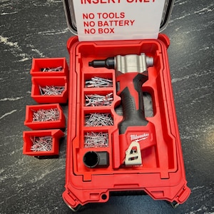 Op de afbeelding: Een rode Milwaukee gereedschapskoffer met een rood-zwarte elektrische gereedschap, zilveren bevestigingsmiddelen en drie rode opbergcontainers. De koffer heeft een label met de tekst "INSERT ONLY. NO TOOLS, NO BATTERY, NO BOX."