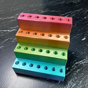 May include: A multi-tiered organizer in a gradient of colors, from pink to blue. Each tier has multiple circular holes, likely for holding pens, pencils, or other small items. The design is modern and colorful.