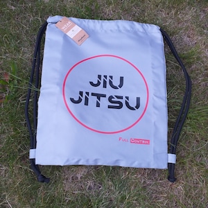 Könnte beinhalten: Eine hellblaue Kordeltasche mit einem roten Kreis und dem Text "JIU JITSU" in Schwarz. Der Text "FULL CONTROL" ist in Rot unter dem Kreis gedruckt.