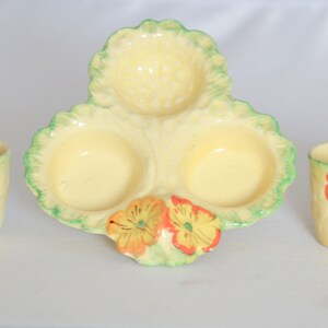 May include: A three-sectioned ceramic egg dish with a yellow glaze and green leaf accents. The dish features three small bowls and is decorated with orange and yellow flowers. Four matching egg cups with a yellow glaze and orange flower accents are also included.