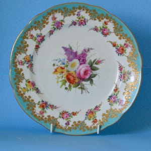 May include: A white porcelain plate with a turquoise rim and gold accents. The plate features a floral bouquet in the center and smaller floral arrangements around the rim. The plate is supported by a white stand.