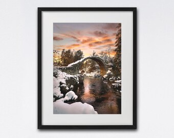 Könnte beinhalten: Eine Steinbogenbrücke über einen Fluss in einem schneebedeckten Wald. Der Himmel ist leuchtend orange und rosa und spiegelt sich im Wasser wider.