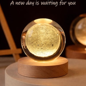 May include: A glowing moon-shaped glass sphere lamp with a wooden base. The lamp is lit from within, illuminating the moon's surface. Text above reads "MAY YOU HAVE A GOOD NIGHT" and "A new day is waiting for you".