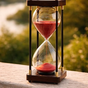May include: An hourglass with a dark wood frame and clear glass bulbs. The sand inside is a vibrant red. The hourglass is supported by black pillars with gold accents. The background is a blurred view of greenery and water.