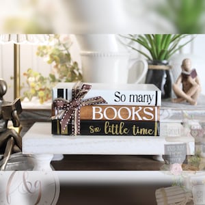 May include: Three stacked decorative books with the text "So many BOOKS so little time" on a white shelf. The books are brown, black, and white with gold accents and a brown ribbon.