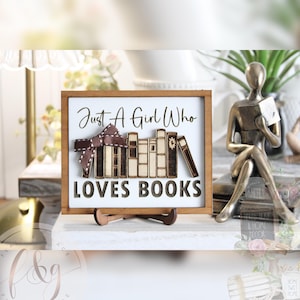 May include: Wooden sign with a white background and a brown frame. The sign reads "Just A Girl Who LOVES BOOKS" in black script and block letters. A decorative bow and wooden books are featured on the sign. The sign is displayed on a wooden stand.
