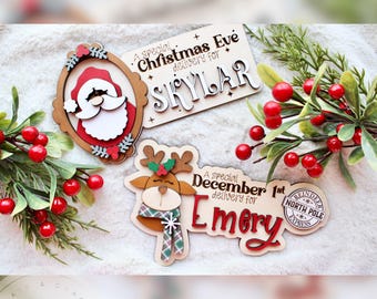May include: Wooden Christmas ornaments featuring Santa Claus, a reindeer, and personalized text. The Santa ornament is framed, while the reindeer has a scarf. The text includes "Christmas Eve delivery for Skylar" and "December 1st delivery for Emery."