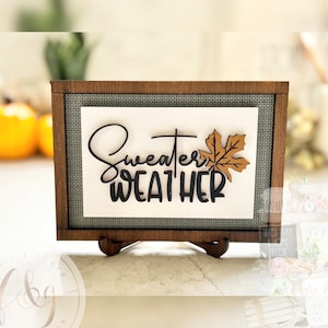 May include: A wooden sign with a grey background and a black and brown leaf design. The sign says "Sweater Weather" in black script.