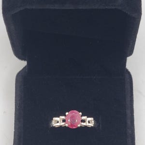 May include: A silver ring featuring a central oval-cut red ruby gemstone held in a four-prong setting. The band has a decorative link-style design on both sides of the stone. The ring is displayed inside a black velvet-lined jewelry box.