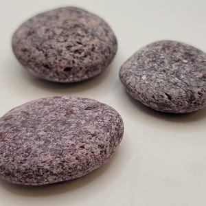 May include: Three smooth, rounded stones with a speckled, textured appearance. The stones have a mix of gray and purple hues, with a slightly rough surface. They are arranged on a white surface.