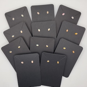 May include: A collection of opal stud earrings displayed on black earring cards. The opals have a range of colors, including white, pink, and yellow. The earrings are presented against a white background, highlighting their colors and details.