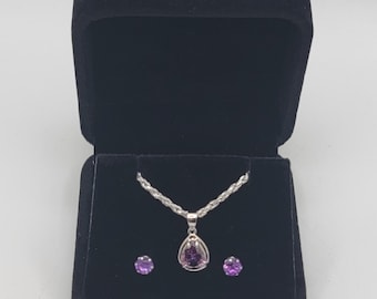 May include: A jewelry set consisting of a silver rope chain necklace with a pear-shaped purple gemstone pendant and two matching round purple gemstone stud earrings, all displayed inside a black velvet-lined jewelry box with a hinged lid.