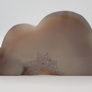 May include: A cloud-shaped agate stone with a polished surface. The stone features a mix of translucent and opaque areas in shades of gray and brown. Small, dark inclusions are visible within the stone, adding texture.