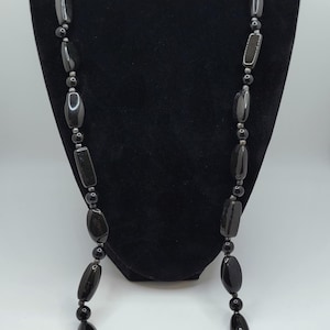 May include: A long, black necklace with various shapes of beads, including ovals, rectangles, and spheres. The necklace is displayed on a black velvet bust against a white background. The beads have a glossy finish.