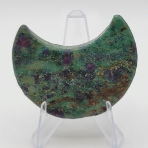 May include: A crescent-shaped gemstone with a green base color and flecks of red and brown. The stone is displayed on a clear plastic stand against a white background. The gemstone has a polished surface.