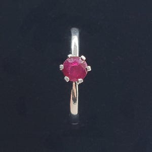 May include: A silver-toned ring featuring a single, round, faceted red gemstone set in a six-prong mounting. The band is smooth and polished, showcasing a simple design that highlights the vibrant red stone at the center of the setting.