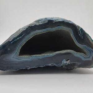 May include: A cross-section of a geode, showcasing a hollow center and layered bands of blue, gray, and white. The outer edge has a rough, textured appearance, while the inner cavity is smooth and dark.