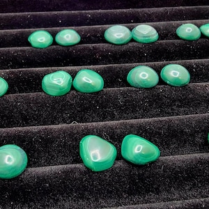May include: A collection of polished malachite stones, each displaying a vibrant green hue with distinctive banded patterns. The stones are arranged on a black velvet surface, showcasing their natural beauty and variations in size and shape.