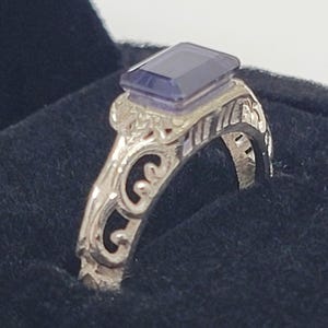 May include: A silver-toned ring featuring an emerald-cut, translucent purple gemstone set atop a band with intricate, openwork scroll patterns. The metal band displays a polished finish with detailed cutouts along the sides of the setting.