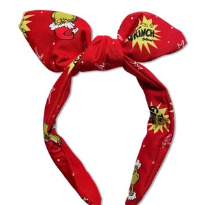 May include: Red headband with a bow and a Grinch print. The headband features the Grinch's face, the words "The Grinch" and "Grinch Dr. Seuss".