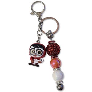 May include: A keychain with a Coco character figurine and a beaded charm. The charm features a large, sparkling red bead, with other beads in shades of orange and white. The keychain has a silver clasp and ring.