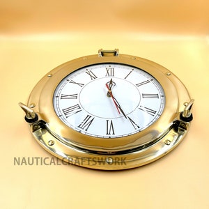 Puede incluir: Un reloj n&aacute;utico de lat&oacute;n con n&uacute;meros romanos en una esfera blanca. El reloj est&aacute; en un marco redondo de estilo ojo de buey con un acabado en lat&oacute;n.