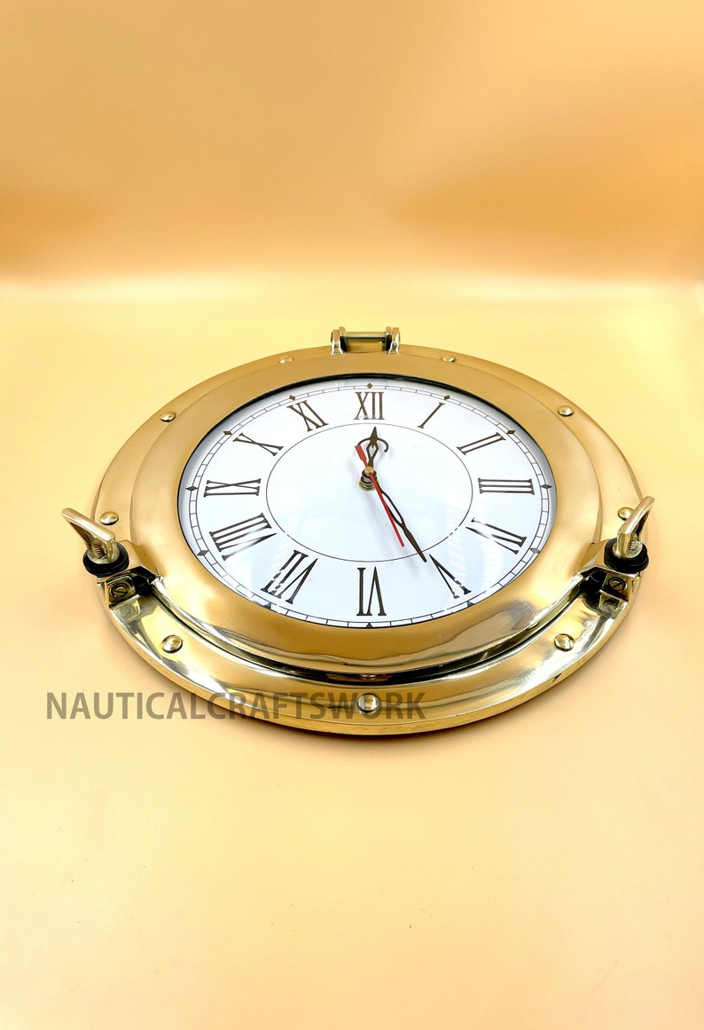 Puede incluir: Un reloj n&aacute;utico de lat&oacute;n con n&uacute;meros romanos en una esfera blanca. El reloj est&aacute; en un marco redondo de estilo ojo de buey con un acabado en lat&oacute;n.
