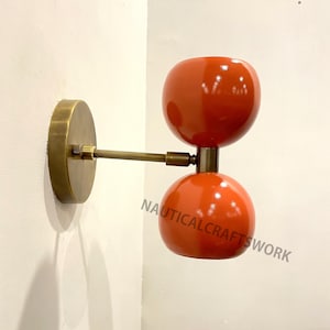 May include: A wall-mounted light fixture with two orange globe-shaped shades and a brass arm. The fixture is mounted on a white wall.