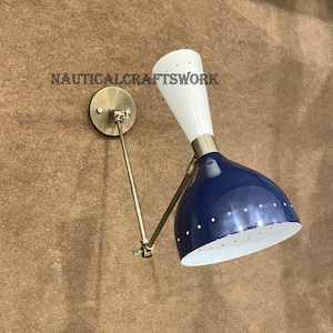 May include: A wall-mounted lamp with a white and navy blue shade, featuring a brass-colored arm and mounting plate. The shade has small white dots and the text "NAUTICAL CRAFTSWORK" is visible.
