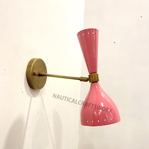 Op de afbeelding: Een roze en messing wandlamp met een uniek ontwerp. De lamp heeft twee lampenkappen, één wijst omhoog en de andere omlaag. De lampenkappen zijn beide roze en hebben een gestippeld patroon. De lamp is gemonteerd op een witte muur.