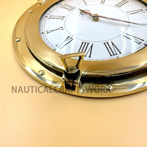 Puede incluir: Un reloj n&aacute;utico de lat&oacute;n con n&uacute;meros romanos y una esfera blanca. El reloj est&aacute; en un marco redondo estilo ojo de buey con un pestillo de lat&oacute;n.