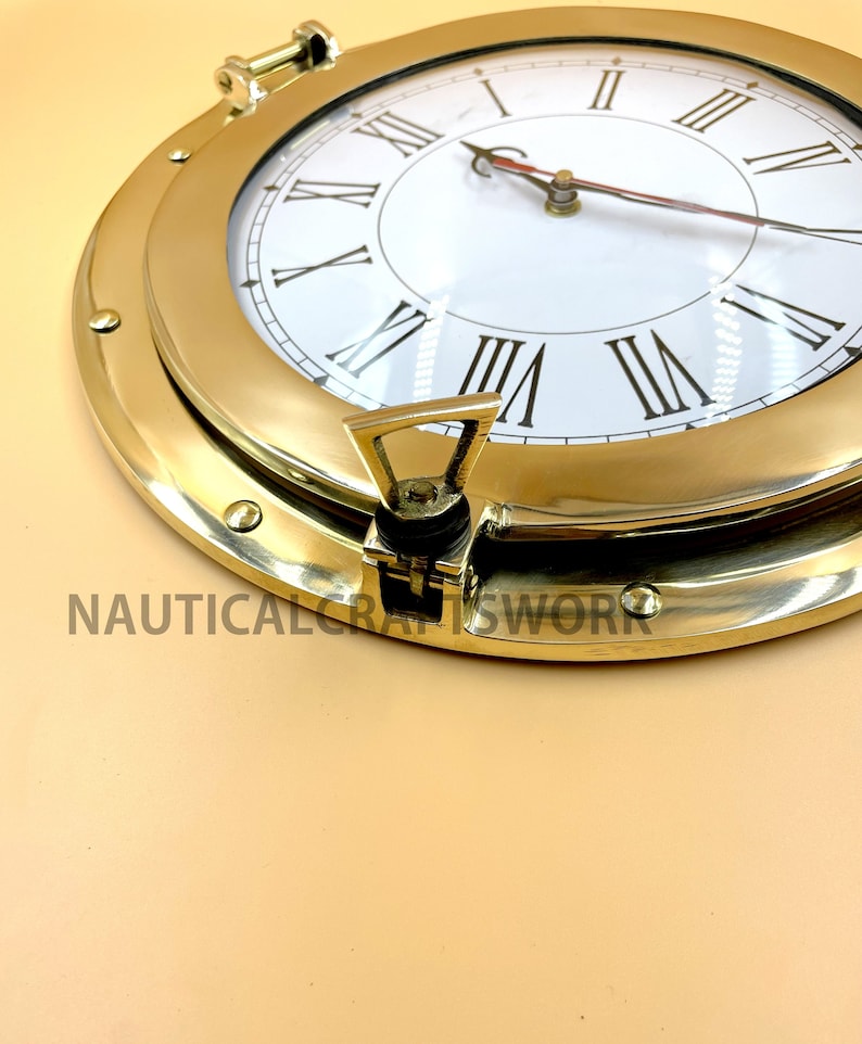 Puede incluir: Un reloj n&aacute;utico de lat&oacute;n con n&uacute;meros romanos y una esfera blanca. El reloj est&aacute; en un marco redondo estilo ojo de buey con un pestillo de lat&oacute;n.