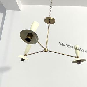 Könnte beinhalten: Ein Messing- und weißer Kronleuchter mit drei Lichtern. Der Kronleuchter hat ein modernes Design mit drei Armen, die sich von einem zentralen Punkt aus erstrecken. Jeder Arm hat einen weißen Schirm mit einer Messingbasis.