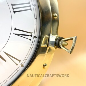 Puede incluir: Un reloj de barco de lat&oacute;n con n&uacute;meros romanos en una esfera blanca. El reloj tiene un bisel de lat&oacute;n y un soporte de montaje de lat&oacute;n. El reloj est&aacute; montado en una pared.