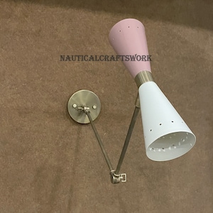 Op de afbeelding: Een messing wandlamp met een witte en roze kegelvormige kap. De kap heeft een patroon van kleine gaatjes. De lamp is verstelbaar en kan omhoog en omlaag worden bewogen.