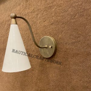 Könnte beinhalten: Eine Wandleuchte aus Messing mit einem weißen, kegelförmigen Schirm. Die Leuchte hat ein schlichtes Design und eine minimalistische Ästhetik.