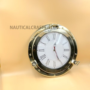 Puede incluir: Un reloj n&aacute;utico de lat&oacute;n con n&uacute;meros romanos en una esfera blanca. El reloj est&aacute; en un marco de ojo de buey redondo de lat&oacute;n con un pestillo de lat&oacute;n.