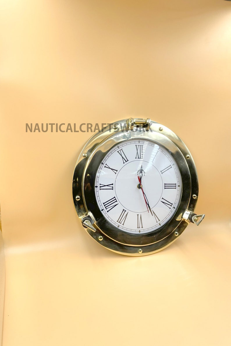 Puede incluir: Un reloj n&aacute;utico de lat&oacute;n con n&uacute;meros romanos en una esfera blanca. El reloj est&aacute; en un marco de ojo de buey redondo de lat&oacute;n con un pestillo de lat&oacute;n.
