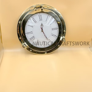 Puede incluir: Un reloj de lat&oacute;n dorado con n&uacute;meros romanos en una esfera blanca. El reloj est&aacute; en un marco redondo de estilo ojo de buey.