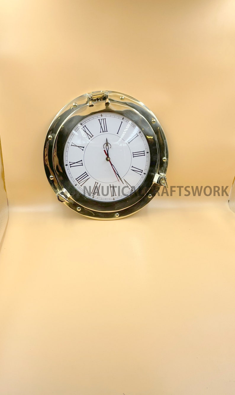 Puede incluir: Un reloj de lat&oacute;n dorado con n&uacute;meros romanos en una esfera blanca. El reloj est&aacute; en un marco redondo de estilo ojo de buey.