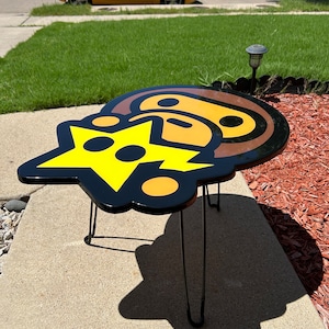 May include: A black and yellow side table with a cartoon character design. The table has three black metal legs.