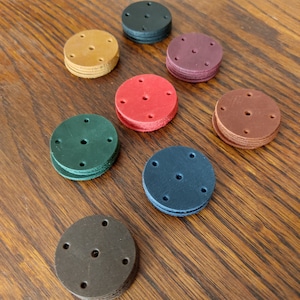 Op de afbeelding: Een set van tien leren schijven in verschillende kleuren, waaronder bruin, rood, groen, blauw en zwart. Elke schijf heeft meerdere gaten die er doorheen zijn geponst.