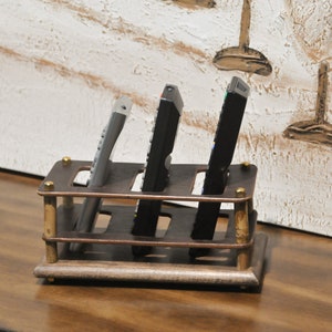 May include: A wooden remote control holder with four slots. The holder is brown with brass accents and is sitting on a wooden surface. Three remotes are in the holder, two black and one silver.