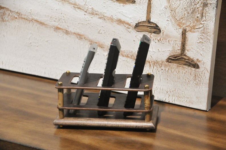 May include: A wooden remote control holder with four slots. The holder is brown with brass accents and is sitting on a wooden surface. Three remotes are in the holder, two black and one silver.