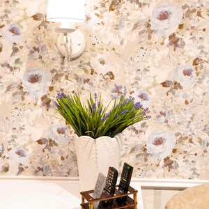 May include: A white wall with a floral wallpaper pattern. A white lamp with a white shade is mounted on the wall. A white vase with lavender flowers sits on a white table. Three remote controls are in a wooden holder on the table.