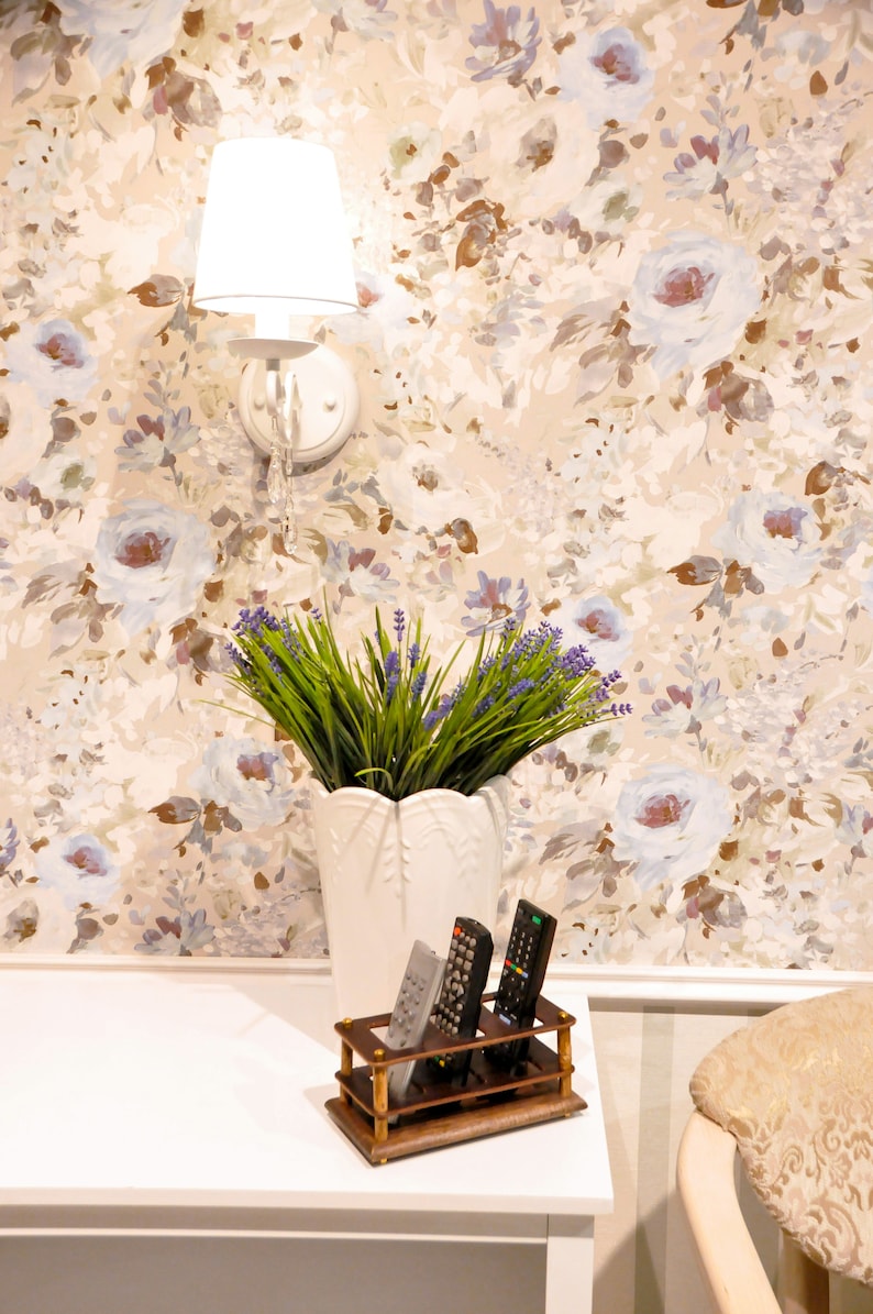 May include: A white wall with a floral wallpaper pattern. A white lamp with a white shade is mounted on the wall. A white vase with lavender flowers sits on a white table. Three remote controls are in a wooden holder on the table.
